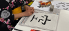A woman in a kimono sits at a table with a piece of paper with the kanji for sky written out